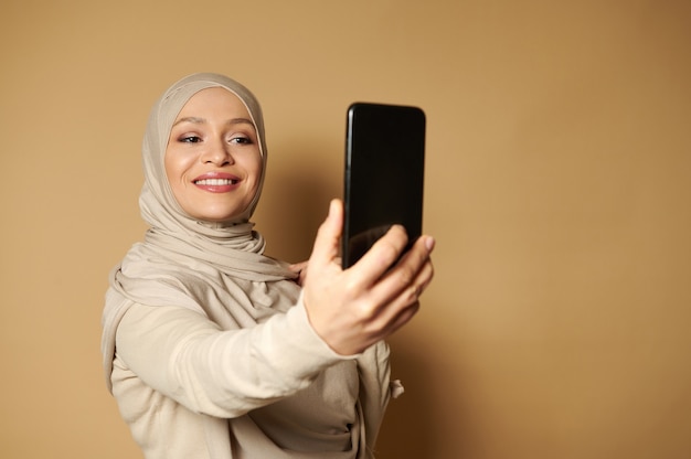 Schöne arabische Frau mit schönem zahnigen Lächeln und bedecktem Kopf, der Selfie macht, das gegen eine beige Oberfläche mit Kopienraum steht