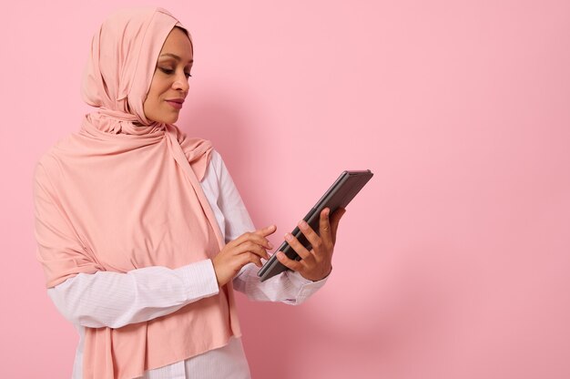 Schöne arabische muslimische Frau mit bedecktem Kopf in rosa Hijab, die vor einem farbigen Hintergrund mit einem digitalen Tablet in den Händen posiert, Kopienraum für Text. Werbung isoliertes Porträt