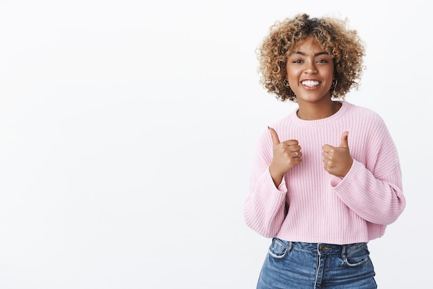 Schöne Arbeit, cooles Ergebnis. Porträt einer freundlich aussehenden, entzückten, attraktiven, fröhlichen Afroamerikanerin mit blondem, lockigem Haarschnitt im Winterpullover, die Daumen zur Unterstützung und Jubel hochhebt, wie