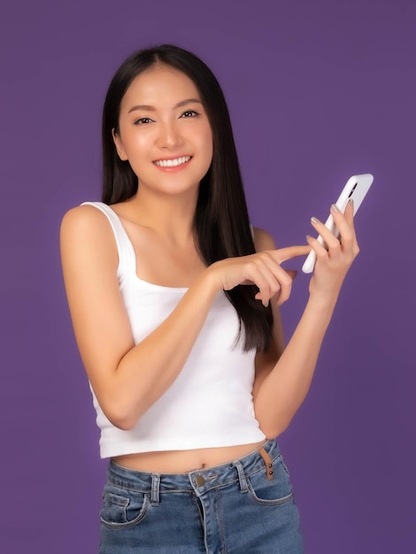 Schöne asiatische brünette Frau, süßes Mädchen in weißem Tanktop, das ein Smartphone spielt, das auf violettem Hintergrund isoliert ist. Mobiltelefon nutzt ein Online-Banking für kostenpflichtiges Online-Shopping