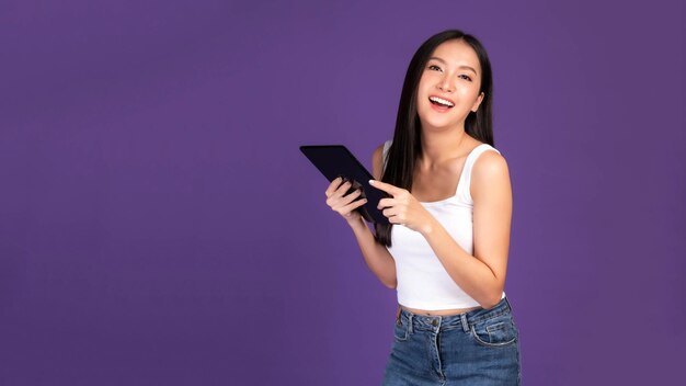 Schöne asiatische brünette Frau, süßes Mädchen in weißem Tanktop, das einen Smartphone-Tablet-Computer spielt, der auf violettem Hintergrund isoliert ist. Mobiltelefon nutzt ein Online-Banking für kostenpflichtiges Online-Shopping