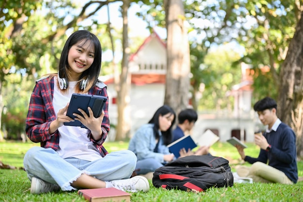 Schöne asiatische College-Studentin, die ihr Tablet benutzt, während sie im Park des Campus sitzt