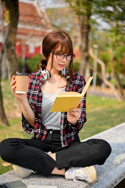 Schöne asiatische College-Studentin, die Kaffee trinkt und sich darauf konzentriert, ein Buch im Park zu lesen