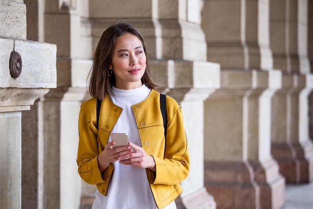 Schöne asiatische Frau benutzt eine Anwendung, um eine SMS-Nachricht auf ihrem Smartphone zu senden. Glückliche junge asiatische Frau mit Smartphone steht auf der Straße.