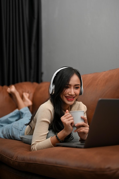 Schöne asiatische Frau, die auf dem Sofa liegt und einen Film auf ihrem Laptop ansieht und Kaffee trinkt