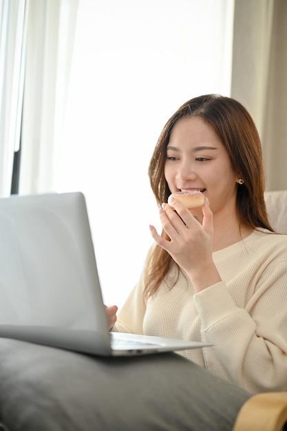 Schöne asiatische Frau, die Donut isst, während sie sich einen Film auf ihrem Laptop im Wohnzimmer ansieht