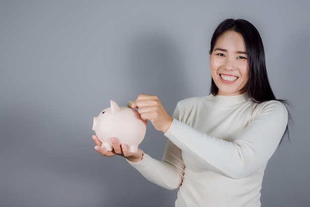 Schöne asiatische Frau, die Münze im Sparschwein auf grauem Hintergrund einsetzt, Kopienraum