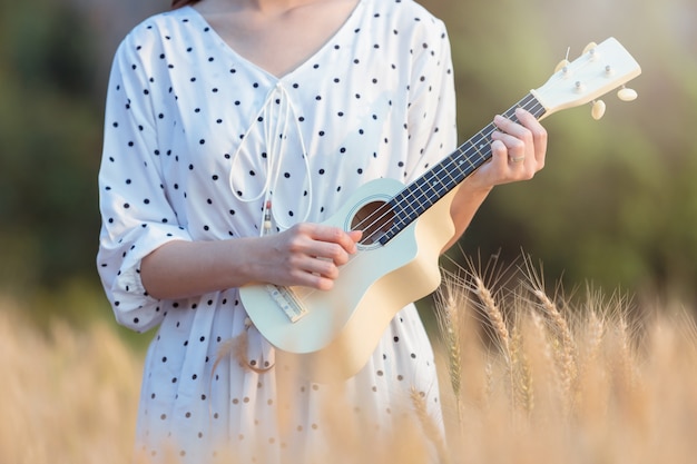 Schöne asiatische Frau, die Ukulele spielt und am Gerstenfeld im Sommer auf Sonnenuntergangzeit sich entspannt