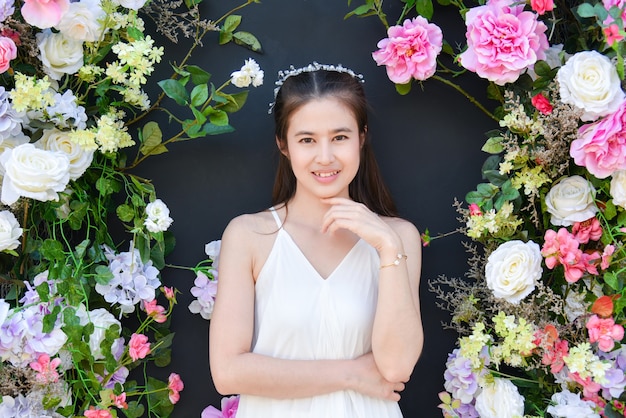 Schöne asiatische Frau, die weißes Kleid trägt, das vor schwarzem Farbhintergrund mit Blume steht.