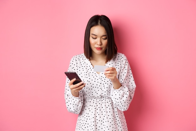Schöne asiatische Frau geben Kreditkarteninformationen in Handy-App ein, kaufen online ein, zahlen für Bestellung auf Smartphone, stehen über rosa.