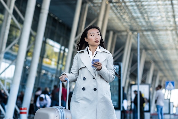 Schöne asiatische Frau, in der Nähe des Flughafens, nutzt eine Telefonanwendung für die Online-Buchung, ein Tourist mit einem großen Koffer