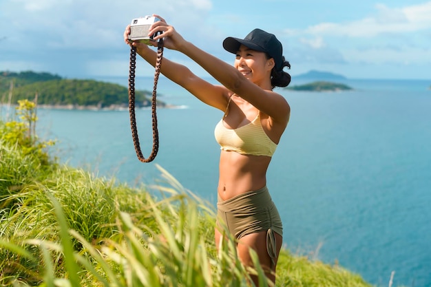 Schöne asiatische Frau in Sportkleidung, die nach dem Trekking ein Foto auf dem Berggipfel am Meer macht