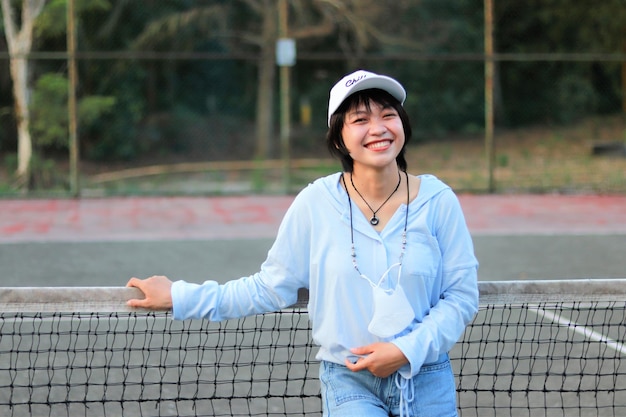 Schöne asiatische Frau mit kurzen Haaren, mit Hut und breitem Lächeln auf dem Tennisplatz