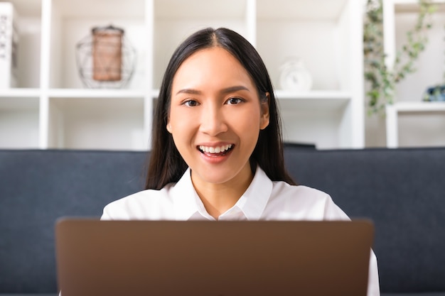 Schöne asiatische Frau mit Laptop und Blick in die Kamera im Wohnzimmer.