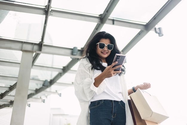 Schöne asiatische Frau mit Sonnenbrille, die ihr Smartphone benutzt und Einkaufstüten hält