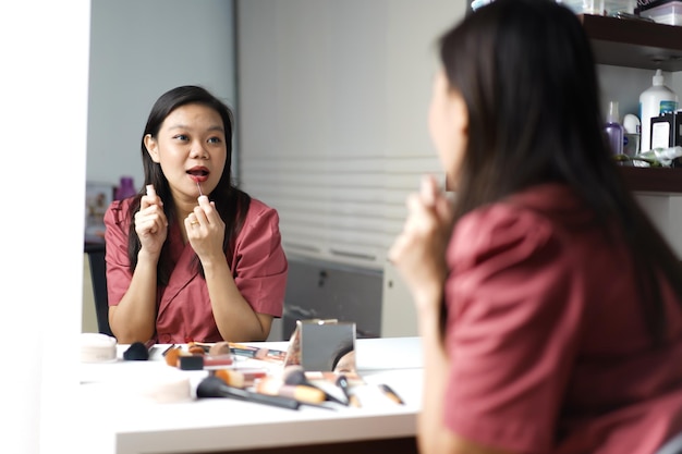 Schöne asiatische Frau schminkt sich und trägt Lippenstift vor einem Schminktischspiegel auf