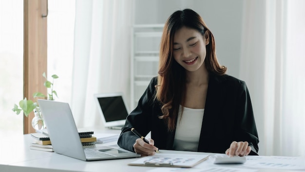 Schöne asiatische Frau sitzt im Büro mit Laptop und Taschenrechner Happy Business Woman sitting an einem Schreibtisch in einem Büro mit einem Tablet-Computer