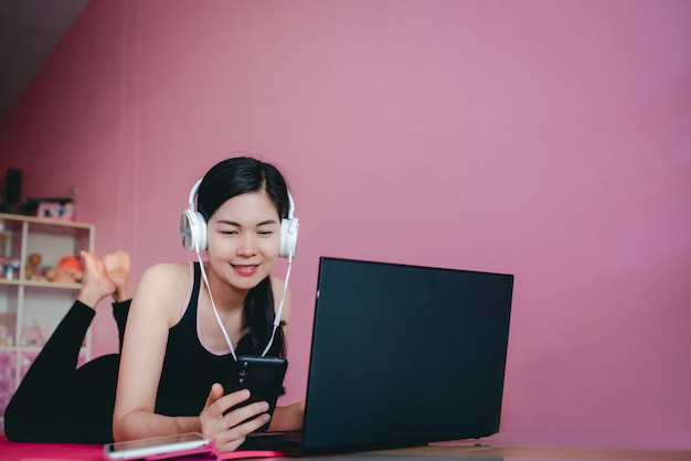 Schöne asiatische Frauen tragen Kopfhörer, trainieren in schwarzer Sportkleidung, legen sich hin, schauen sich Heimtrainingsvideos und Notebooks an.