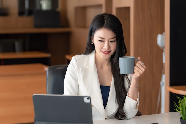 Schöne asiatische Geschäftsfrau, die eine Kaffeetasse hält, die mit einer Tablette im Büro arbeitet.