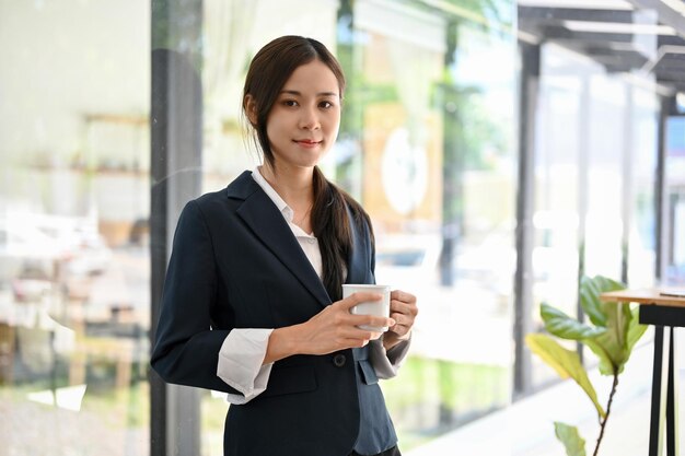 Schöne asiatische Geschäftsfrau, die mit einer Tasse Kaffee in ihren Händen vor ihrem Büro steht