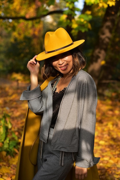 Schöne asiatische Geschäftsfrau im grauen Anzug, gelbem Hut, gelbem Mantel im Park in der Nähe des Sees Autumn