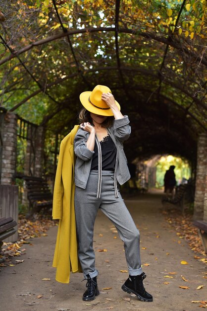 Schöne asiatische Geschäftsfrau im grauen Anzug, gelber Hut hält einen gelben Mantel in ihren Händen im Park Herbst