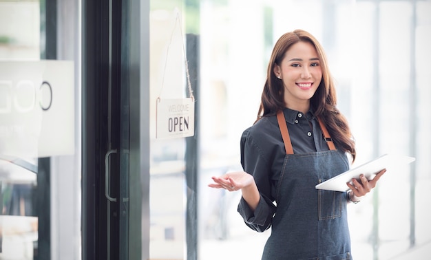 Schöne asiatische junge Barista-Frau in Schürze, die Tablette hält und vor der Tür des Cafés mit offenem Schild steht. Startup-Konzept für Geschäftsinhaber.