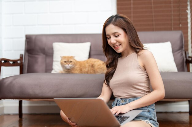 Schöne asiatische junge Frau benutzt Laptop, während ihre Katze zu Hause auf dem Sofa im Wohnzimmer sitzt