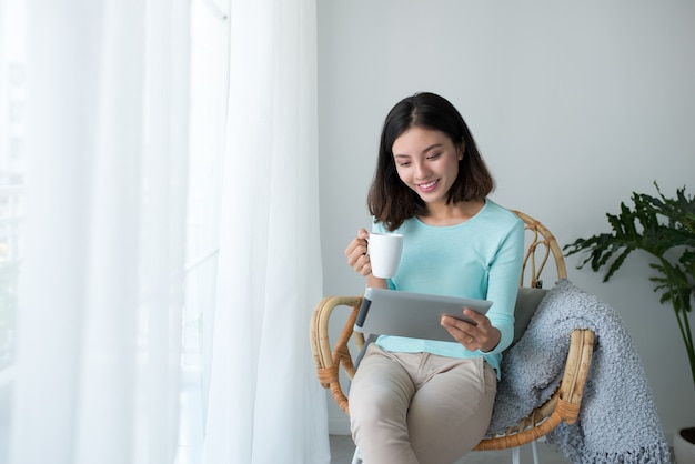 Schöne asiatische junge Frau, die sich zu Hause entspannt und digitale Tablette verwendet