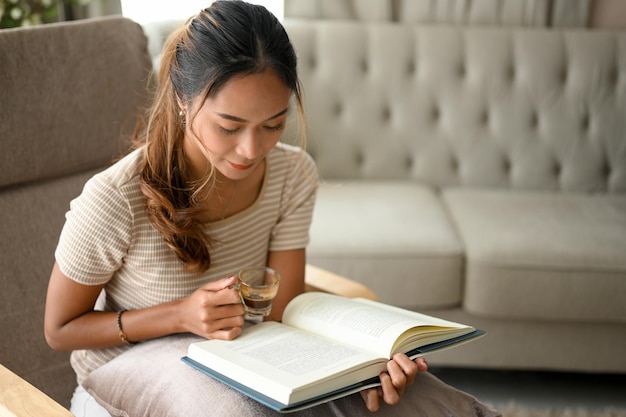 Schöne asiatische junge Frau in lässigem Outfit, die ein Buch in ihrem modernen Wohnzimmer liest Hobby-Konzept