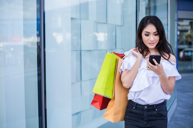 Schöne asiatische junge Frau mit Einkaufstaschen unter Verwendung ihres Smartphones mit Lächeln beim Stehen am Bekleidungsgeschäft. Glück, Konsum, Verkauf und Menschenkonzept