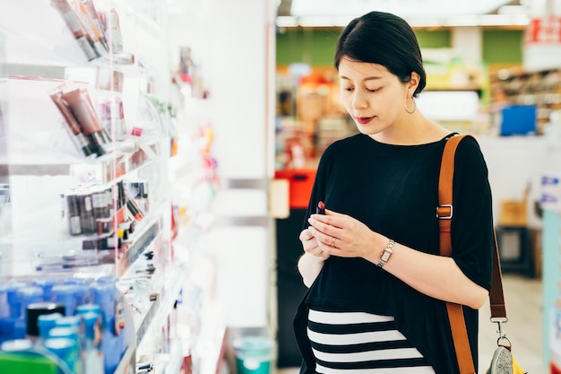 Schöne asiatische koreanische schwangere Frau, die kosmetische Produkte im Supermarkt kauft. charmante junge zukünftige mutterschaftsdame mit dickem bauch, der make-up-lippenstifte hält und farbe im lebensmittelgeschäft auswählt.