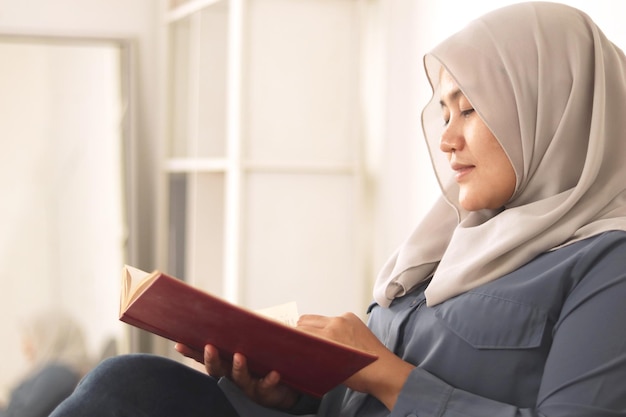 Schöne asiatische muslimische Frau liest ein Buch, während sie auf dem Sofa sitzt.