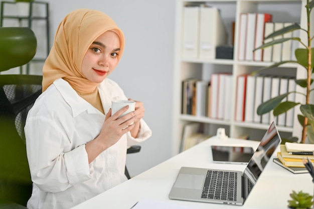 Schöne asiatische muslimische Geschäftsfrau mit einer Tasse Kaffee sitzt an ihrem Schreibtisch im Büro