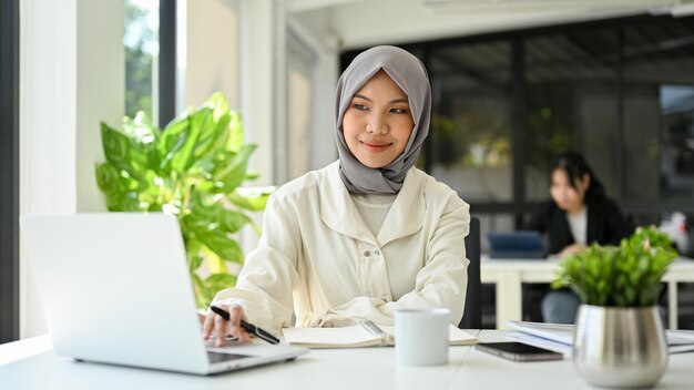 Schöne asiatische muslimische Geschäftsfrau mit Hijab und Laptop im Büro