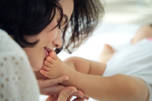 Schöne asiatische Mutter, die ihre Babyfüße mit schließend Augen küsst, während das Kind schläft