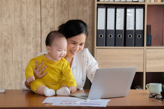 Foto schöne asiatische mutter verbrachte zeit mit neugier kleinkind baby reden, spielen am arbeitsplatz zu hause, entzückenden ungezogenen sohn glücklich lachen mit mutter halten in der hand, alleinerziehende mutter füttert multitasking arbeiten