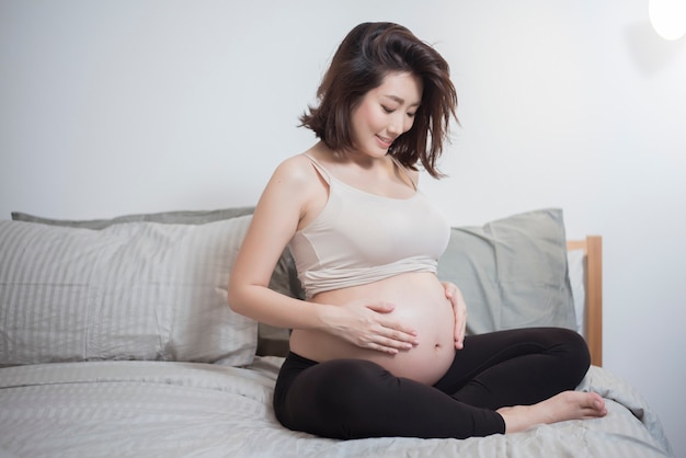 Schöne asiatische schwangere Frau sitzen auf Bett und berühren ihren Bauch