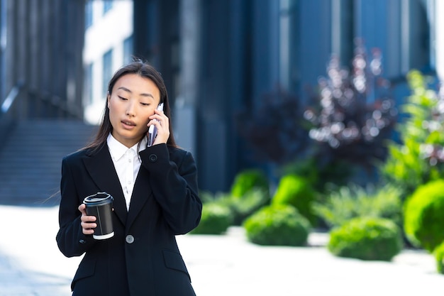 Schöne asiatische Studentin, die telefoniert, während sie in der Nähe des Universitätscampus spazieren geht, eine Frau, die eine Tasse heißes Getränk hält, berichtet über die Neuigkeit der Zulassung