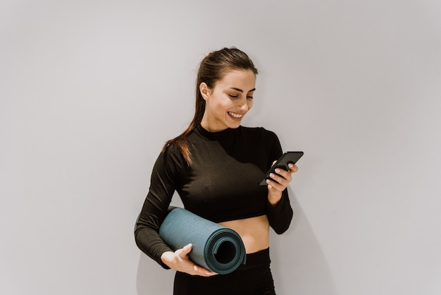 Schöne athletische Frau, die ein Mobiltelefon verwendet und eine Yogamatte hält