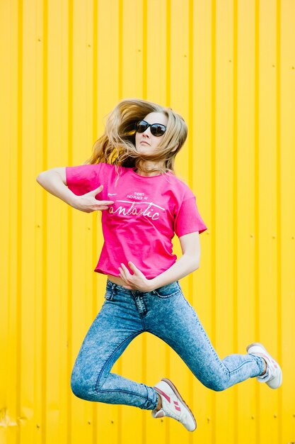 Schöne athletische junge Frau mit langen blonden Haaren im rosa Hemd, blaue Jeans. In weißen Turnschuhen. Posieren, lächelnd an der Wand der Garage an der gelben Wand Platz für Text. Brille. fliegend. springt