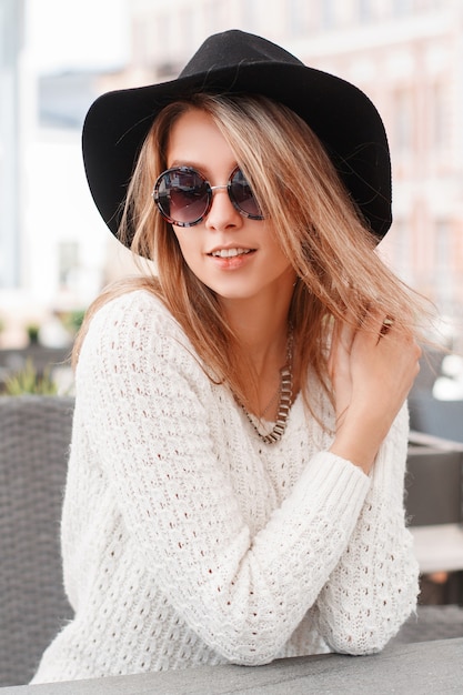 Schöne attraktive junge Frau in einem weißen Vintage-Strickpullover in einem eleganten Hut in modischer Sonnenbrille ruht an einem warmen Frühlingstag in einem Straßencafé. Charmantes freudiges blondes Mädchen.