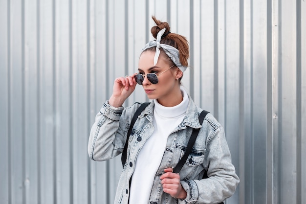 Schöne attraktive junge Hipster-Frau in einem stilvollen Kopftuch mit einer stilvollen Frisur in modischer Sonnenbrille in einer Sommer-Jeansjacke mit einem Rucksack nahe der Metallwand