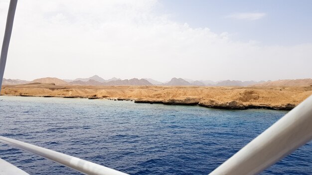 Schöne Aussicht vom Deck eines Kreuzfahrtschiffes im Roten Meer in Ägypten. Ägyptische felsige Küstenlandschaft mit einer Yacht. Ein Teil des Schiffes vor der Kulisse der Wüste und des Meeres.