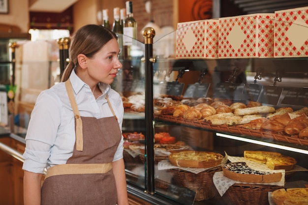 Schöne Bäckerin, die an ihrem Bäckereigeschäft geht und Gebäck auf dem Display betrachtet, kopiert Raum