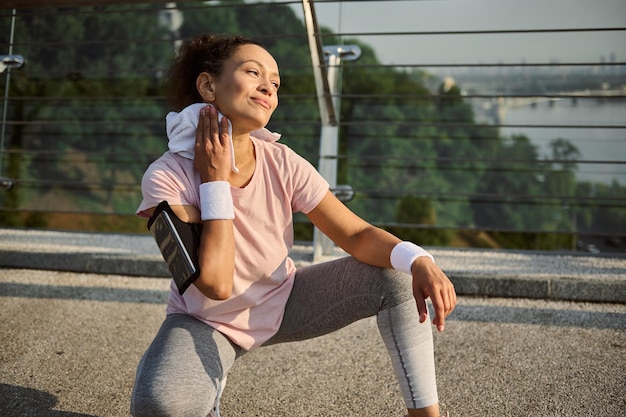 Schöne begeisterte hispanische Sportlerin, die sich mit weißem Frotteetuch den Hals vom Schweiß abwischt, auf der Stadtbrücke in der Hocke sitzt, sich nach dem Joggen entspannt und den sonnigen Sommertag während des Trainings genießt