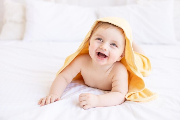 Schöne blonde Baby mit einem gelben Handtuch auf dem Kopf nach einem Bad oder Baden auf einem weißen Bett zu Hause in einem hellen Raum lächelt das Konzept der Kinderpflege und Hygiene