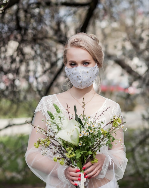 Schöne blonde Braut in einem Hochzeitskleid und einer medizinischen Schutzmaske auf ihrem Gesicht,