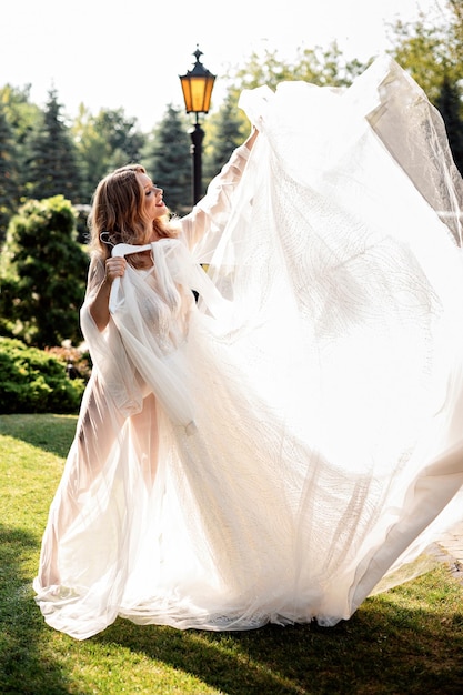 Schöne blonde Braut in weißem Kleid hält mit ihrem stilvollen Hochzeitskleid einen Hängemann, glückliche junge Frau posiert an der frischen Luft, sonniges Wetter in der grünen Natur.