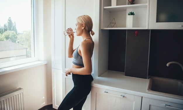 Schöne blonde Frau, die ein Glas Wasser trägt, das Sportkleidung zu Hause trägt